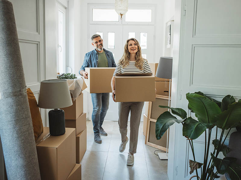 2 people carrying boxes through a house with boxes and items stacked up.